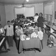 A group of approximately twenty-eight young children of varying ages, and one teacher, stand in a George Washington Carver Day Nursery classroom. They are in fancy dress and are around a table with punch and treats. One boy salutes the camera.