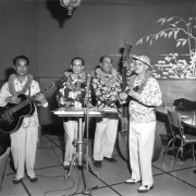 Interior photograph of four musicians playing music (possibly) at the Lotus Lounge for the Chinese New Year Celebration. Each of the musicians wears a floral shirt; they play the guitar, keyboard, cello, and the ukulele.