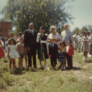 Outdoor group photograph with (L-R) Burnis McCloud, Lillian S. Bondurant, Dorothy Reaves, Elvin Caldwell, and eight children. Ms. Bondurant holds a gold shovel with a green ribbon to celebrate the new George Washington Carver Day Nursery location at 2270 North Humboldt Street in the CIty Park West neighborhood. The yard where they stand was eventually built into the school's new playground. Additional people are seen in the background that are also attending the celebration.