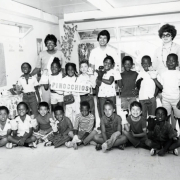 An indoor, group photograph of students and teachers at the George Washington Carver Day Nursery located on 2270 N. Humboldt in the City Park West neighborhood. The Carver Nursery divided their students into classes based on age group, and bestowed names, such as "Kidettes" and "Tom Thumbs," on each class in the school. This group, named the "Pinocchios" included sixteen students and three teachers. The children hold a sign reading "Pinocchios" and hand puppets. 