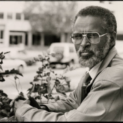 Tsegaye Hailu, who immigrated to the United States from Ethiopia, is now a hydrogeologist at J.F. Sato's engineering firm. He sits with his hands clasped facing the camera. He is wearing sunglasses.