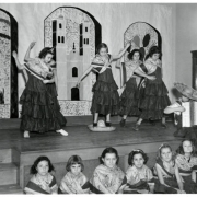 Photograph of several older female students from Ebert Elementary School performing a hat dance.  Five girls stand on stage dressed in traditional Mexican clothing.  They have a sombrero on the floor between them.  They are all in the middle of dancing, while an unidentified boy sits on the stage with an accordion and wearing a sombrero.  Several other girls dressed in the same costumes sit on the floor in front of the stage.