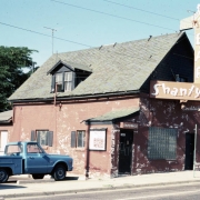View of the Shanty Bar at 151 South Broadway in the Baker neighborhood of Denver, Colorado. The modified residential structure has a gabled roof with a dormer window. There is a covered entrance and a large vertical electric neon sign that reads: "Shanty Bar."  Graffiti marked on sign on the side of the building reads "Barb Menudo."  The structure was built around 1900.