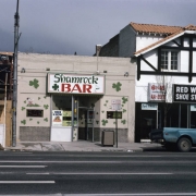 View of the Shamrock Bar at 54 South Broadway in the Speer neighborhood of Denver, Colorado. This one story brick commercial style building has modified windows and a recessed entry way. The brick has been painted beige with green shamrocks, a lighted sign above the door is decorated with a shamrock and a champagne glass and reads: "Shamrock Bar." The Red Wing Shoe Store is on the south side of the building and a construction site is to the north. The structure was built in 1907.
