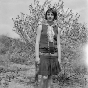 A woman poses in front of a gnarled bush outside Keota, Colorado in Weld County. She wears a flapper-style dress sleeveless bodice and pleated skirt; her hair is in a bob.
