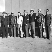 Chief of Internal Security, Tomlinson, and a group of Japanese police staff pose in a line in front of "Block 6F", Camp Amache, Granada Relocation Center, southeastern Colorado. Tomlinson wears a suit, vest, tie and hat, his staff wear jackets or sweaters, some have hats and eye glasses. All wear arm bands designating their rank, left to right, Patrolman, Desk Sergeant, Sergeant, Lieutenant, Captain, and Chief. The Chief holds a cigarette.