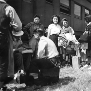 Japanese evacuees stand or sit with their suitcases and belongings in front of a Santa Fe and Topeka passenger train car. The men and women wait for the bus ride to Camp Amache, Granada Relocation Center, southeastern Colorado.