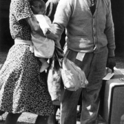 View of a Japanese family on their way to the Granada Relocation Center, Camp Amache, Prowers County, southeastern Colorado, includes two women in dresses, heels and bonnets. One holds a baby, and a man in a sweater, tie, eye glasses and a hat, holds a suitcase. The man has a tag on his belt and another on the suitcase.