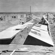 Men hoist a section of prefabricated wall at the Granada Relocation Center, Camp Amache, Prowers County, southeastern Colorado. Other army-style barracks show in various states of construction; some with roofs, others just with framing and composite board walls. Prairie is in the background.