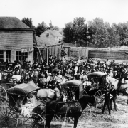 A crowd in Canon City, Colorado, gathers for the laying of the cornerstone of the Mount Moriah Masonic Lodge, at the southeast corner of 5th (Fifth) and Main. People carry umbrellas and cornets, and wear uniforms, sack suits and hats. Horses and buggies are in the foreground, and frame buildings in the background include the Chinese laundry, with its projecting sign reading: Laundry.