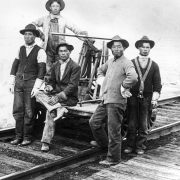 Five Japanese workers pose around a railroad handcart. Each wears  workclothes, including heavy boots, hats, and gloves.