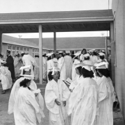 Young Japanese women in white graduation gowns, high heels and mortar board caps and a men in dark gowns and caps assemble in a courtyard at the Granada Relocation Center, Camp Amache, Prowers County, southeastern Colorado. The wind blows their gowns and the women's curled hair. Some read commencement programs, some stand under a covered walkway.