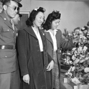 Two Japanese soldiers in uniform pose with their girlfriends by a flower exhibit, at the Arts and Crafts Festival, Granada Relocation Center, Camp Amache, Prowers County, southeastern Colorado. The women wear long wool coats, white blouses, skirts, and ribbons in their curled hair. One holds an artificial flower in the display. The soldiers wear glasses and sunglasses.