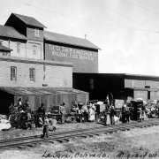 Migrant workers gather with their children along the tracks of the Denver &amp; Rio Grande railroad in La Jara, Colorado in Conejos County. Their possessions, including bedframes, cabinets, wardrobes, tables, chairs, and kitchen utensils are gathereed at the side of the tracks.  The La Jara Milling and Elevator Co. is in the background.  Signs: "The La Jara Milling and Elevator Co. Wholesale Flour Grain and Feed."