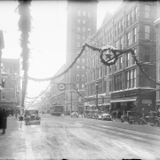 A snowy 16th (Sixteenth) street, Denver, Colorado, is decorated for Christmas with garlands of evergreen and wreathed stars that hangs over the street. The Daniels and Fisher Tower at Arapahoe Street is on the right. In the foreground the marquee for the Tabor Theater reads: " "[Ta]bor [?] Her Life [?] S Moran [?] Pickford [?] al Comedy [?] Li fe [?] oran."; signs read: "Ford's Glasses Kodaks." Shows automobiles, trolley cars and trolley lines.