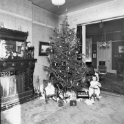 Interior view of a living room in a house in (probably) Denver, Colorado; shows small children and toys by a decorated Christmas tree. Shows: fireplace, toy piano, stuffed animals, and doll.