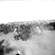 Native American men and women take part in a Bear Dance near Ignacio, La Plata County, Colorado.  A line of men face a line of women while groups of white and Native American men, some standing near a wooden pole and brush fence, all wearing hats, look on.