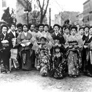 A group of Japanese-American women and young girls dressed in kimonos pose in the middle of a street, possibly in Denver, Colorado. Some of the women hold miniature American flags. A man stands next to the group; he wears a suit, tie and hat. Front row (behind young girls), first from left is Mrs. Reverend Ono; second from left is Mrs. N. Hokasono. Front row third from right is Mrs. Y. Hayano and second row, first from right is Mrs. M. Otsuki.