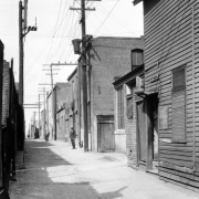 View of Hop Alley (between Wazee and Blake Streets) in Denver, Colorado. Men who include a Chinese man and a police officer stand near the brick buildings. A sign reads: "1417 Wing Gut Clothing Co." and has Chinese characters.