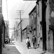 Men walk on Hop Alley (between Wazee and Blake Streets) in Denver, Colorado. Another group of men sit near an automobile.