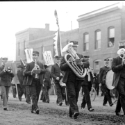 View of a marching band in a funeral procession probably on Wazee Street in Denver, Colorado. They play instruments that include: baritones, trombones, clarinets, and trumpets. Buildings line the street.