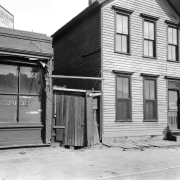 A man poses near a two-story wood frame building on either Wazee or Blake Street near Hop Alley in Denver, Colorado. Signs on nearby buildings and windows read: "2032", "Conway Bogue Realty Inv. Co. Real Estate In All Branches Fire Insurance 524 17th St", and "Public Station Local And Long Distance Telephone".