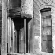 View of the entrance to a building on Hop Alley in Denver, Colorado. The wooden door has a padlock and screened window. The address numerals read: "2051".