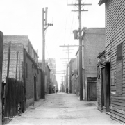 Men who include a police officer stand in Hop Alley (between Wazee and Blake Streets) in Denver, Colorado. A sign with Chinese characters is on a wood frame building.
