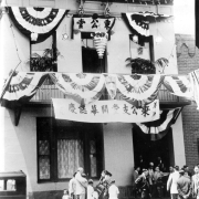 Chinese men, women, and children stand outdoors near the Chinese Masonic Lodge Number 185 possibly at 1926 Market Street in Denver, Colorado. The two story building is decorated with bunting, paper lanterns, and banners with Chinese characters.