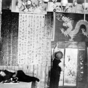 A Chinese-American man hangs a scroll with Chinese characters on a wall in a building on Hop Alley in Denver, Colorado. Celestial lays nearby and smokes possibly opium from a water pipe. Images of a dragon and flowers are on the wall.