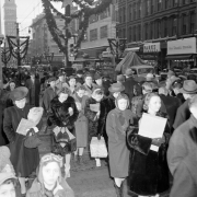 Men, women, and children stand on the sidewalk near 16th (Sixteenth) Street in downtown Denver, Colorado. Nearby streetlights are decorated with greenery for Christmas.