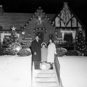 Charles Lilburn Cousins, an African American, poses with his wife and daughter in front of their house at 2525 York Street, at night.  The house and trees in the yard are decorated for Christmas and New Year's with garland, ornaments, stars, and signs that read: "Noel," "Merry Christm[as]," and "Happy New Year."