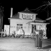 Sarah Sims, an African-American woman, wears a choir robe, holds a book, and stands in front of her house at night.  The house is decorated for Christmas.  Statues and cut outs of choir singers stand in a line in front of the house.  A large paper birthday cake is decorated with electric candles and has numbers on each of the 3 layers: "1000," "900," "57."  A sign at the top of the house reads: "Our Savior's Birthday."  Additional signs are: "Dec. 25th," and "Peace on Earth."