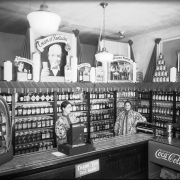 Chinese women and an African-American man stand behind the counter of a liquor store in Denver, Colorado. Liquor bottles and displays read: "Cream of Kentucky," "Old Quaker," "Wilken Family," "Coca-Cola," and "Golden Wedding".