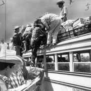 A man checks a roster as Mexican migrant workers climb on a bus owned by the Great Western Sugar Company in Colorado. Luggage and passengers are on the roof of the bus.