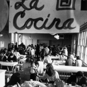 Hispanic and white men, women, and children eat and wait in line for food in a migrant worker camp in Colorado. A hand-made sign reads: "La Cocina."