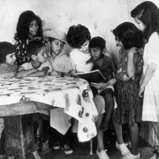 A white woman reads to Hispanic boys and girls at a migrant laborer camp near Greeley (Weld County), Colorado. One boy wears a wide-brimmed hat. The girls wear dresses.