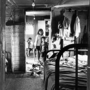 Interior view of a migrant worker's house in Colorado. A bed, clothes, and radio are in one room. Boys and a girl sit and stand in a nearby room.