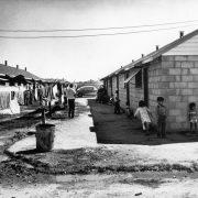 Boys, girls, and a man stand near laundry on a clothesline near Fort Lupton (Weld County), Colorado. One-story cinder block houses with pitched roofs are nearby. An automobile is parked in the distance.
