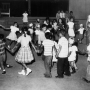 Hispanic women, boys, and girls dance at a birthday party in a migrant worker's camp near Fort Lupton (Weld County), Colorado. The women and girls wear dresses. One boy wears shorts.