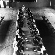 Migrant workers eat at a long table in a gymnasium in Colorado. A cook stands at the end of the table. A basketball hoop is in the room.