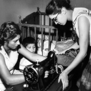 A Hispanic woman sews with a sewing machine in Colorado. A boy and white woman look on. A baby crib is nearby.