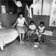A Hispanic woman sits on a bed with her children at her house in Colorado. A doll and jacket are on the beds. Curtains and clothes on hangers hang from the walls nearby.