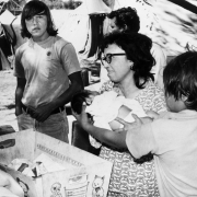 A Mexican American woman holds a baby at a migrant worker camp in Colorado. Boys and a man stand nearby. Tents are in the distance.