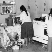 Women cook in the kitchen of a migrant worker's house in Colorado. Pots, pans, a stove, and a wooden crate used as a shelf are in the room.