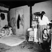 A woman cooks in a migrant worker's house near Brighton (Adams County), Colorado. A small boy stands nearby and holds a baby bottle. Pots, pans, a pail, stove, mattress, and bunk beds are in the room.