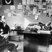 James Fresques, State Social Services Director Con F. Shea, John Jones, and Migrants in Action members Leonel Sanchez and Jim Mason meet in a Migrants in Action office in Colorado. A television, space heater, and desk are in the office. Banners, newspaper clippings, and signs are on the walls.