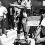 Robert Valenzuela, a Mexican American and Director of the Colorado Migrant Council, gives a speech during a protest in Colorado. Other protesters stand nearby. One man holds a sign that reads: "Colorado Migrant Council Is Unfair Unjust &amp; It Stinks!"