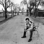 A Hispanic migrant worker sits on a chair in road probably near Fort Lupton (Weld County), Colorado. Migrant workers' houses line the dirt street.