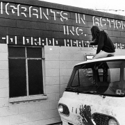 A woman paints a sign on the side of a building during a protest over migrant workers' living conditions probably near Fort Lupton (Weld County), Colorado. She sits on the roof of a van. The sign reads: "Migrants in Action Inc. Colorado Headquarters."
